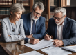 smart middle aged man and woman sat at a table with a smart looking lawyer all looking at documents